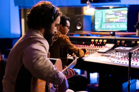 Songwriter creating a new song with his guitar in control room, playing electro acoustic instrument and singing next to record producer. Young artist performing live, editing music on mixing console.