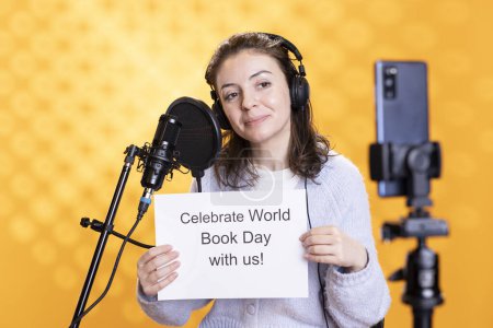 Aufmunternde Person mit Plakat mit Botschaft zum Welttag des Buches, Filmen mit Telefon und Mikrofon, Studiohintergrund. Strahlender Bücherwurm nimmt Video auf, das Lese- und Schreibkompetenz fördert