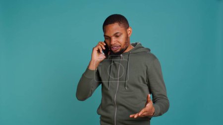 Irritierter afrikanisch-amerikanischer Mann, der sich während eines Telefonats mit Freunden streitet, isoliert über Studiohintergrund. Empörte Person streitet mit Kumpels während eines Gesprächs auf Handy, Kamera B