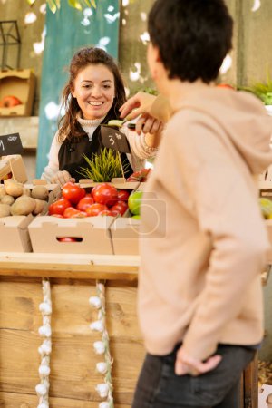 Smiling merchant gives female client fruit sample for tasting and selling healthy at local farmers market. Friendly woman farm stand owner offering consumer to try eco products before purchasing it.