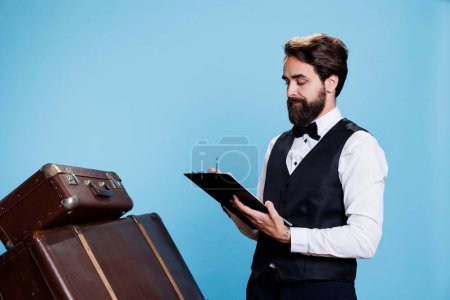 Téléchargez les photos : Hotel concierge takes notes on papers to keep evidence of guests, verifying bulletpoints on log register. Employee in formal attire reading and reviewing archive records of reservations in studio. - en image libre de droit