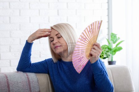Mujer madura que experimenta sofocos de la menopausia en casa. Esta foto captura la incomodidad de los sofocos durante la menopausia, mientras una mujer lucha por enfriarse con un delicado ventilador de papel..