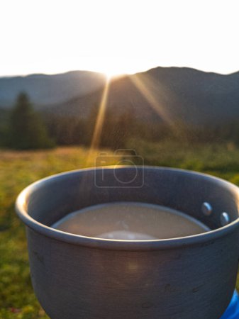Foto de Primer plano del café de la mañana con leche en una taza de senderismo contra el telón de fondo del sol naciente en las montañas - Imagen libre de derechos