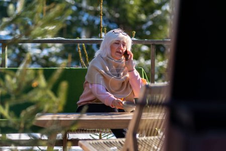 Foto de Una mujer habla por teléfono mientras está sentada en la terraza de su casa de campo y bebe café. - Imagen libre de derechos