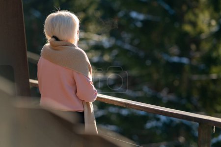 Foto de Una mujer está de pie en el balcón con su espalda a la cámara y mira hacia el bosque de invierno. - Imagen libre de derechos