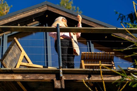 Foto de Mujer sosteniendo teléfono inteligente mientras está de pie en la terraza de su casa entre los muebles de exterior - Imagen libre de derechos