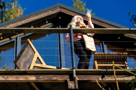 Foto de Una mujer adulta mira el teléfono mientras está de pie en la terraza de su casa con una bufanda tirada sobre sus hombros. - Imagen libre de derechos