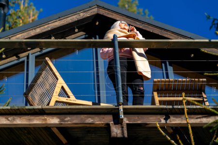 Foto de Vista inferior de una mujer hablando por teléfono, de pie en la terraza de su casa en un bosque de coníferas - Imagen libre de derechos
