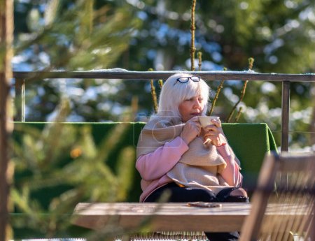 Foto de Una mujer bebe café o té mientras está sentada en una mesa en la terraza de su casa en el bosque. - Imagen libre de derechos