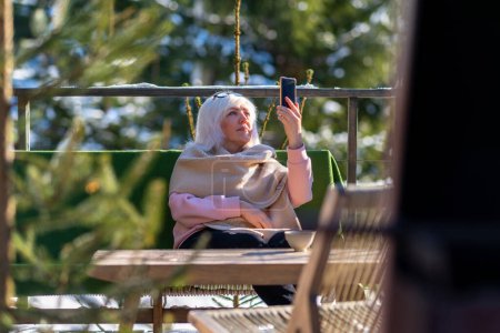 Foto de Una señora adulta se comunica por vídeo en el teléfono mientras está sentada en una silla cubierta con una manta en la terraza de su casa en un día soleado de invierno. - Imagen libre de derechos