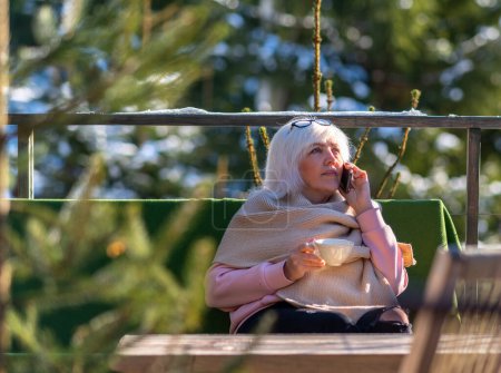 Foto de Una señora está hablando por teléfono, sentada en una mesa en la terraza y tomando café o té.. - Imagen libre de derechos