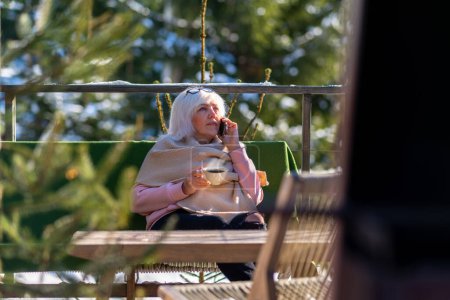Foto de Una mujer bebe una taza de café mientras está sentada en la terraza de su casa con una bufanda tirada sobre sus hombros en un cálido día de invierno.. - Imagen libre de derechos