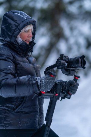 A woman in winter clothes holds a stabilizer with both hands and watches the video recording process.