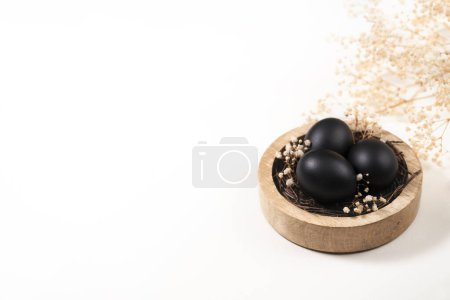 Photo for Eggs painted black on white background. Nest containing three egg with beautiful gypsophila flowers. Minimal Easter concept with copy space for text. - Royalty Free Image