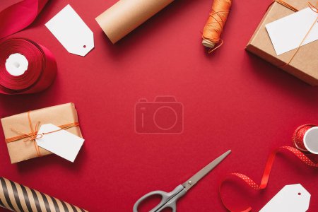 Photo for Kraft gift boxes with orange thread on a red background. Packaging and preparation of gifts for the celebration. Christmas New Year backdrop with scissors, paper and labels for inscriptions - Royalty Free Image