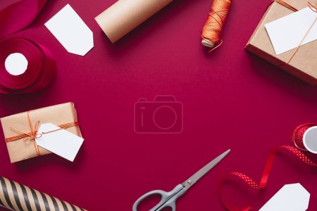 Photo for Kraft gift boxes with orange thread on a red background. Packaging and preparation of gifts for the celebration. Christmas New Year backdrop with scissors, paper and labels for inscriptions - Royalty Free Image