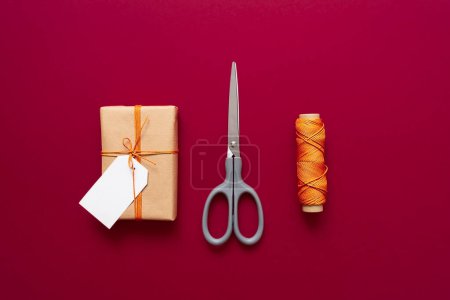 Téléchargez les photos : Coffret cadeau Kraft avec fil orange, ciseaux et étiquettes pour inscriptions sur fond rouge. Emballage et préparation des cadeaux pour la célébration. Noël Nouvel An toile de fond - en image libre de droit