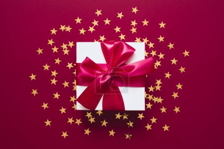 Téléchargez les photos : Coffret cadeau blanc avec ruban sur un rouge avec des confettis dorés en forme d'étoiles. Valentines, Noël ou Nouvel An en toile de fond. Style de pose plat avec un design minimaliste. - en image libre de droit