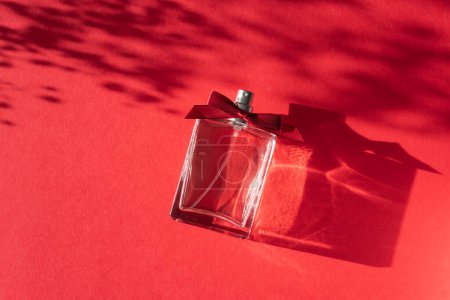 Téléchargez les photos : Transparent bottle of perfume with ribbon bow on a red background. Fragrance presentation with daylight. Trending concept in natural materials with shadows. Womens essence.Valentines Day Gift. - en image libre de droit