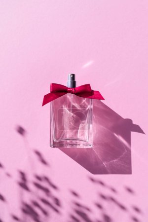Téléchargez les photos : Valentines Day Gift. Transparent bottle of perfume with ribbon bow on light pastel colored lilac background. Fragrance presentation with daylight. Trending concept in natural materials with shadows. - en image libre de droit