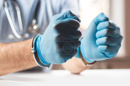A medical officer, doctor, or quack in a blue medical uniform in handcuffs. The concept of crimes committed by medical personnel or providing medical services without a license.