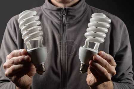 Téléchargez les photos : Concept d'économie d'énergie. Main masculine tenant deux ampoules sur fond sombre. - en image libre de droit