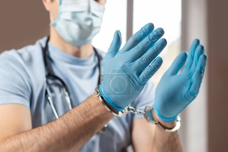 A medical officer, doctor, or quack in a blue medical uniform in handcuffs. The concept of crimes committed by medical personnel or providing medical services without a license.