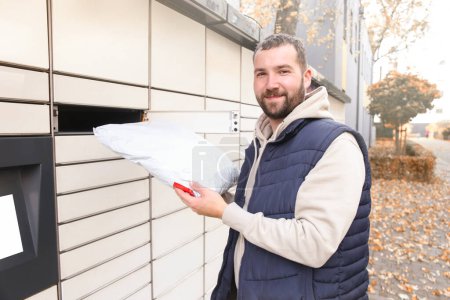 Photo for Happy bearded man put the parcel in the self-service mail terminal. Parcel delivery machine. - Royalty Free Image