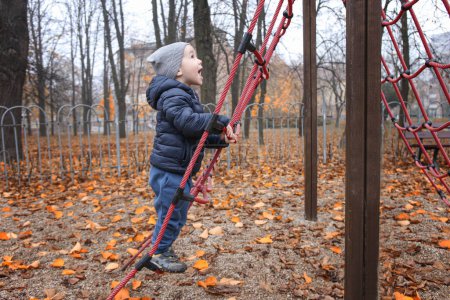 Foto de Un niño de 2 años juega en el patio de recreo en otoño, un niño feliz trepa cuerdas deportivas en la estación fría y juega - Imagen libre de derechos