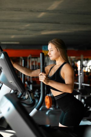 Portrait of luxurious young woman with blonde hair is engaged in gym on an orbitrek elliptical trainer. Young blonde girl with sexy figure doing cardio workout.