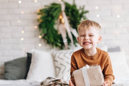Photo for Cute happy Caucasian boy opening his presents on Christmas morning. Christmas tree wreath on background. Child excited and smiling. Happy family holidays at home. Copy space. High quality photo - Royalty Free Image