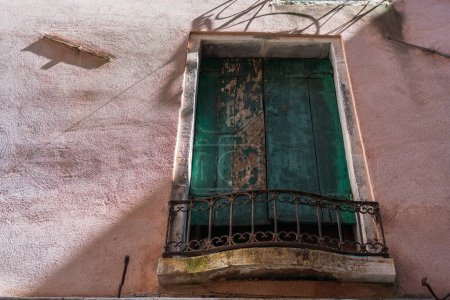 Foto de Una gran ventana vintage con viejas persianas de madera verde en una pared áspera en la ciudad de Venecia, Italia, un rayo oblicuo de luz solar en la pared de una casa antigua, un juego de sombra y luz - Imagen libre de derechos