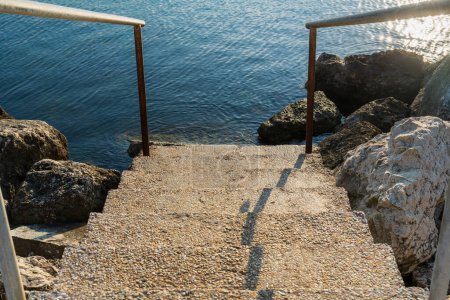 Photo for Stone steps and metal railings leading to the sea, morning sunlight, beautiful water surface, large stones on the seashore, romantic simple path, photorealistic natural background, postminimalism - Royalty Free Image