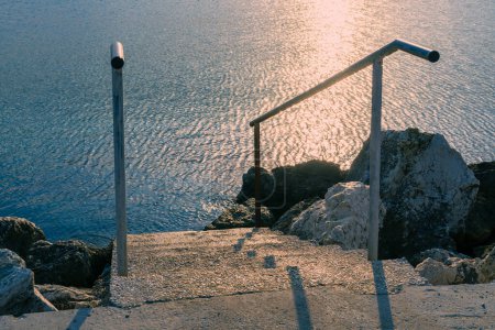 Photo for Tone stairs and metal railings lead to the sea, morning sunlight on the surface of the blue sea, large stones on the shore, romantic path, photorealistic natural background, post-minimalism - Royalty Free Image