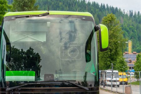 Foto de Primer plano de un gran parabrisas de un autobús turístico en una parada de autobús, fondo borroso de árboles y autobuses verdes, reflejos de nubes y árboles en el cristal del autobús, viajes de verano - Imagen libre de derechos