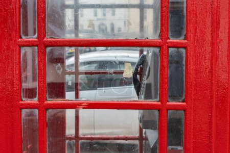 Foto de Primer plano de la icónica cabina telefónica roja británica con teléfono público en el interior y ventanas de cristal. Vidrio sucio. Escena urbana callejera con coche y edificio en segundo plano. - Imagen libre de derechos