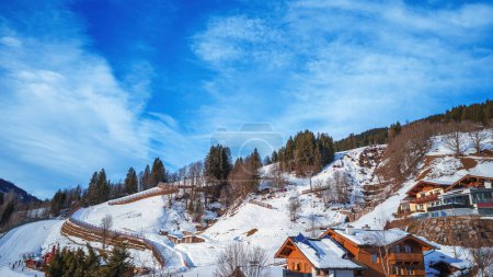 Eine ruhige Winterlandschaft mit gemütlichen Holzhütten schmiegt sich an einen schneebedeckten Hügel, unter einem strahlend blauen Himmel mit wehenden Wolken. Ideal für Konzepte der Ruhe, des Urlaubs und des alpinen Lebens