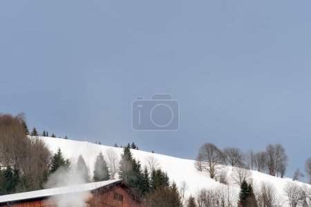 Photo for A serene winter landscape, showcasing a snow-covered hill with trees and a cabin emitting smoke, under a clear blue sky. Ideal for concepts of tranquility, winter retreats, and natural beauty - Royalty Free Image