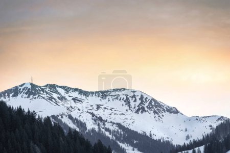 Ein ruhiger Blick auf einen schneebedeckten Berg bei Sonnenuntergang, der das Konzept der natürlichen Schönheit und Ruhe verkörpert. Der goldene Himmel steht im Kontrast zu den dunklen, bewaldeten Ausläufern und bietet einen Moment der Besinnung