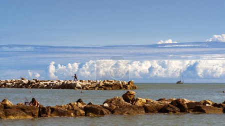 Serene seascape with people on a rocky beach, a distant sailboat, and majestic clouds, capturing the essence of peaceful outdoor relaxation