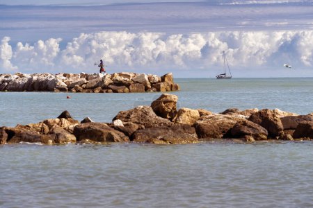 Un pescador solitario se encuentra a orillas rocosas, arrojándose al mar con un velero y nubes encima, encarnando la paz y la belleza de la naturaleza.