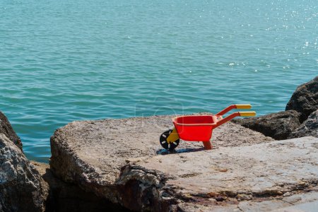 Eine rote Spielzeugschubkarre an einem felsigen Ufer vor glitzernder Meereskulisse weckt Kindheitserinnerungen und die Freude am Spiel am Meer