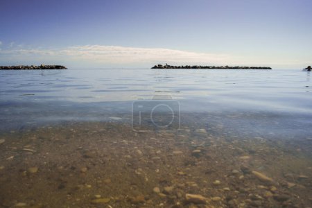 Serene waterscape with pebbled bottom, calm surface, distant rock barrier under clear skies ideal for peaceful imagery