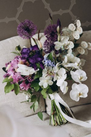 Foto de Wedding Floristics. Bridal bouquet of pink, white and purple flowers and greenery with white ribbon lies on the couch in the room - Imagen libre de derechos