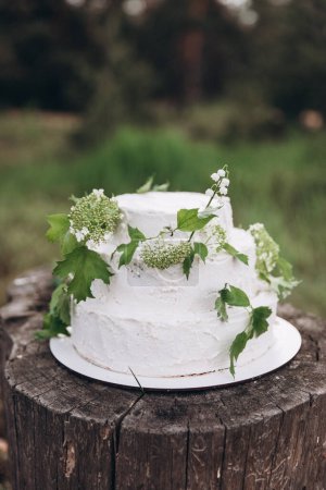 Foto de Pastel de boda blanco de tres niveles hecho de crema y galletas se encuentra en un tocón en un bosque de verano y decorado con ramas de vegetación y flores - Imagen libre de derechos