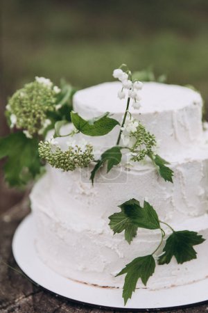 Téléchargez les photos : Gâteau de mariage blanc à trois niveaux fait de crème et de biscuit se dresse sur une souche dans une forêt d'été et décoré avec des branches de verdure et de fleurs - en image libre de droit