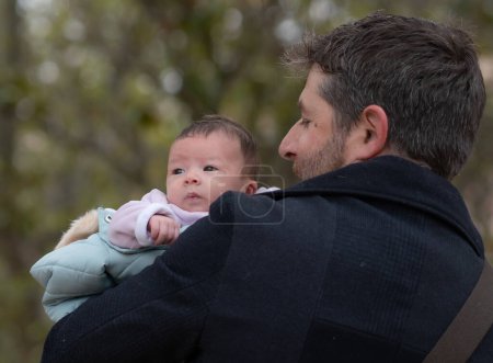 Photo for Outdoors lifestyle portrait of happy father holding his newborn baby girl only a few weeks old sitting on city park bench taking care of the little daughter proud and cheerful - Royalty Free Image