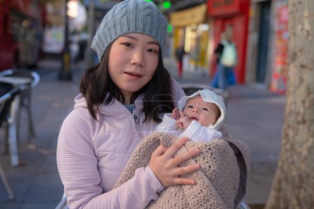 Téléchargez les photos : Portrait de mode de vie urbain extérieur de jeune femme chinoise asiatique heureuse et belle comme mère de petite adorable petite fille tenant sa tendre et douce dans la rue de la ville le jour ensoleillé d'hiver - en image libre de droit
