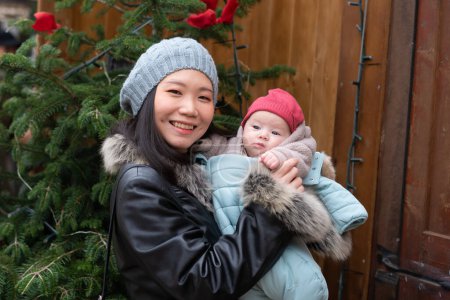 Photo for Young happy and attractive Asian Chinese woman holding baby girl in her arms in front of Christmas tree walking on Winter street market in mother and daughter love concept - Royalty Free Image
