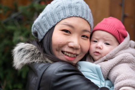 Téléchargez les photos : Jeune femme chinoise asiatique heureuse et attrayante tenant bébé fille dans ses bras devant l'arbre de Noël marchant sur le marché de rue d'hiver dans le concept d'amour mère et fille - en image libre de droit